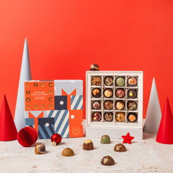 A festive chocolate collection gift set displayed on a white surface against a solid red-orange background. The set includes a colorful patterned box labeled 'FESTIVE COLLECTION', an open white box filled with assorted chocolates, and several loose chocolates scattered in front.