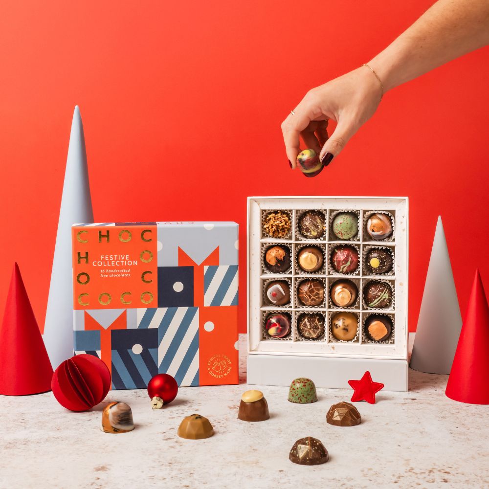 A hand reaches for a chocolate from an open white box of assorted chocolates, with a Chococo branded gift box beside it and several chocolates scattered on a light surface against a bright red background.